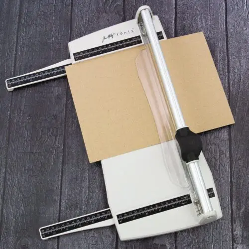 The Tim Holtz Rotary Media Trimmer has a light grey plastic base with grid marks debossed on the surface. It has a imperial and metric rulers with an extendable base and a geared rotary trimmer arm. In this frame, there is a piece of kraft cardstock on the trimmer, ready to cut. The extendable arms are out to cater for a slightly larger cardstock. The image is on a faux wooden background.
