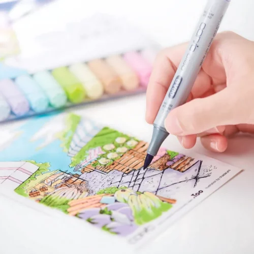 close up of someone using a copic ciao marker and writing on a paper and colouring in a picture of a garden. there is a set of copics in the background.