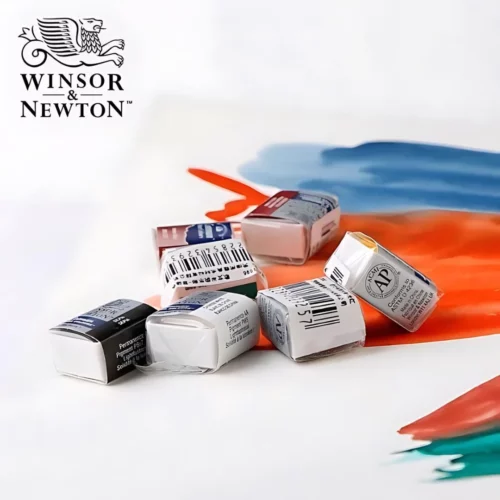 A promotional image for Winsor and Newton Cotman Half Pans. There are a pile of half pans laying in the center of the frame on a piece of paper that has been painted with watercolour paint in blue, orange and a small amount of green. The winsor and newton logo is shown in the top left hand corner of the frame.