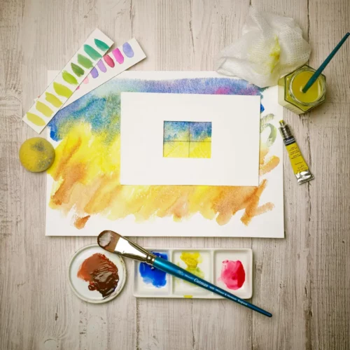 A promotional image for Winsor and Newton Cotman Watercolour Tubes. It is a birds eye view of a piece of paper on a wooden table top which has been paint with the Cotman paints. There is a tube of yellow paint to the right of the paper and a glass jar with a paint brush in the jar above the tube of paint. Below the picture is a palette with a paint brush laying on top of it.