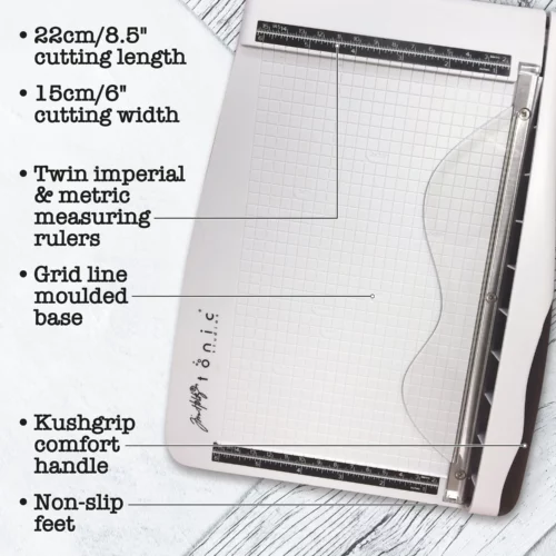 A promotional image for the Tim Holtz Guillotine Trimmer. The trimmer has a light grey plastic base and a guillotine arm with a black kushgrip handle for comfort. The Tim Holtz logo is shown in the image. On a light grey background.