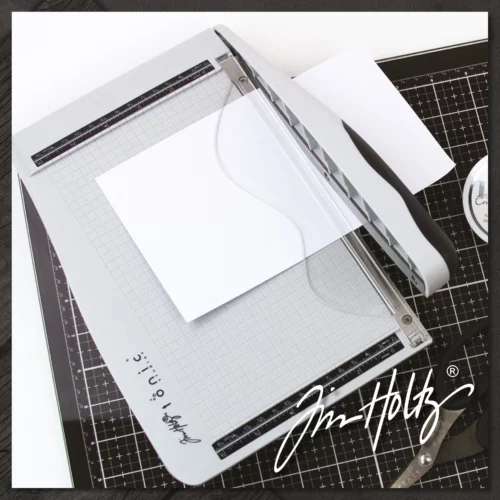 A promotional image for the Tim Holtz Guillotine Trimmer. The trimmer has a light grey plastic base and a guillotine arm with a black kushgrip handle for comfort. The Tim Holtz logo is shown in the image. On a dark grey background.