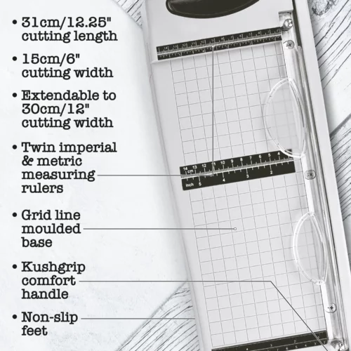 A promotional image for the Tim Holtz Maxi Guillotine Comfort Paper Trimmer. The trimmer has a light grey plastic base and a guillotine arm with a black kushgrip handle for comfort. The Tim Holtz logo is shown in the image. On a light grey background.