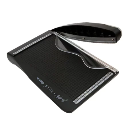 The Tim Holtz Deckle Torn Edge Trimmer has a black plastic base with grid marks debossed on the surface. It has a guillotine arm that is also black and clear plastic paper guide over the top of the trimmer. The trimmer is rectangular in shape and is shown in the frame at a slight angle on a white background.