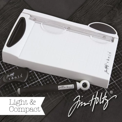 A promotional image for the Tim Holtz MINI Trimmer. The trimmer has a light grey plastic base and a guillotine arm with a black kushgrip handle for comfort. The Tim Holtz logo is shown in the image. On a dark grey background.