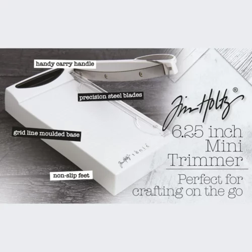 A promotional image for the Tim Holtz MINI Trimmer. The trimmer has a light grey plastic base and a guillotine arm with a black kushgrip handle for comfort. The Tim Holtz logo is shown in the image.