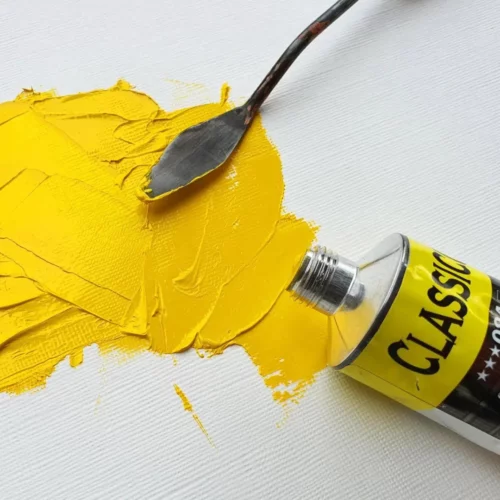 There is a single tube of Maimeri Classico Oil Paint laying on it's side, coming in from the right hand side of the frame. The tube is open and there is yellow paint on a surface in front of the tube, with a palette knife seen coming out of the top of the frame, laying on the yellow paint. The image is a close up and is cut off by the frame.