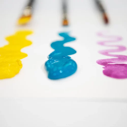 A promotional image for Van Gogh Watercolours. There are 3 samples of paint on a white surface. Yellow on the left, blue in the middle and pink on the right. Behind each pile of paint is a paint brush but it is blurred in the background.