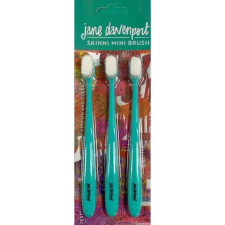 The Jane Davenport Skinni Mini Brush Set of 3 is shown in the center of the frame. The set has a printed cardboard backing that is printed with the Jane Davenport logo. The brushes are held to the cardboard backing with a thick clear plastic. You can see the 3 brushes through the clear packaging. The brushes have a teal handle and white bristles and resemble a toothbrush. On a white background.