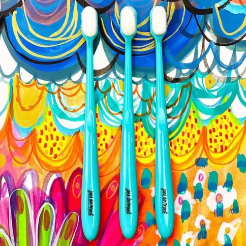 The brushes from the Jane Davenport Skinni Mini Brush Set are shown loose in the frame on a background that was made using the brushes and paint. The brushes have a teal handle and white bristles and resemble a toothbrush. On a white background.