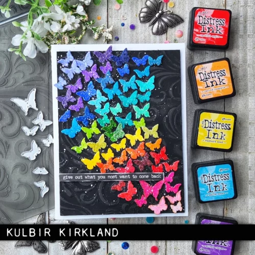 A card is shown in the frame that was made using the Vault Boutique by Tim Holtz Thinlits Die Set. The card front is fille with tiny little butterfly cutouts that are rainbow coloured and appear to be flying off the page.