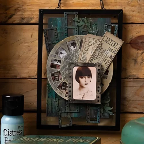 An card was is shown in the frame which was made by using the Vault Picture Show by Tim Holtz Thinlits Die Set. The card front is shown with a picture wheel and some filmstrips behind them and an old photo is attached to the top of the elements. In sepia and brown tones. A vintage style card.