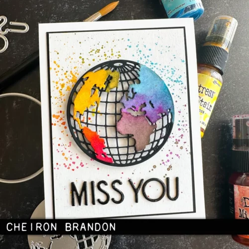 A greeting card was made using the Vault World Travel by Tim Holtz Thinlits Die Set and is shown in the frame. The card front has a world map on the front which is brightly coloured and the words 'miss you'.