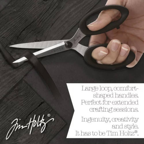 A promotional image for Tim Holtz Haberdashery Scissors. A persons hand is seen coming in from the right hand side of the frame, using the scissors to cut a piece of ribbon. On a faux wooden background. The Tim Holtz logo is shown in the bottom left hand corner of the frame.