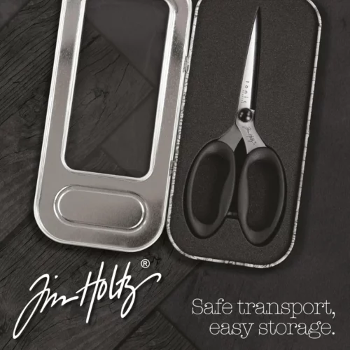 A promotional image for Tim Holtz Haberdashery Scissors. The scissors are inside the tin and the tins lid is open. Laying on a faux wooden background and the Tim Holtz Logo is shown in the bottom left hand corner of the frame.