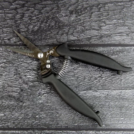 A pair of Tim Holtz Mini Recoil Snips can be seen laying on a faux wooden background. The snips have a black plastic handle and a metal blade with a spring between the handles. Laying on a faux wooden background.