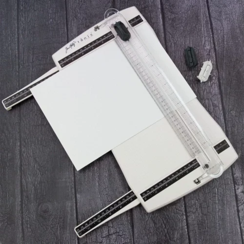 The Tim Holtz Precision Trimmer has a light grey plastic base with grid marks debossed on the surface. It has a imperial and metric rulers with an extendable base and a geared rotary trimmer arm. In this frame, there is a piece of white cardstock on the trimmer, ready to cut. The extendable arms are out to cater for a slightly larger cardstock. The image is on a faux wooden background.