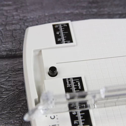The Tim Holtz Precision Trimmer has a light grey plastic base with grid marks debossed on the surface. It has a imperial and metric rulers with an extendable base and a geared rotary trimmer arm. In this frame, a close up of the top of the trimmer is shown. There is a black line with imperial and metric markings printed on it. You can see the top of the rotary arm in this frame. On a faux wooden background.