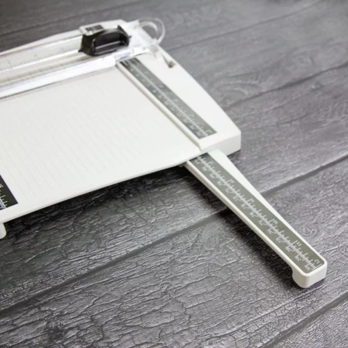 The Tim Holtz Precision Trimmer has a light grey plastic base with grid marks debossed on the surface. It has a imperial and metric rulers with an extendable base and a geared rotary trimmer arm. In this frame, a close up of the corner of the trimmer is shown coming in from the top right hand side of the frame with one of the extendable rulers shown coming out of the trimmer. The image is on a faux wooden background.