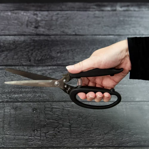 A persons hand is seen coming out of the right hand side of the frame holding a pair of Tim Holtz Recoil Scissors. The scissor handle is black. There is a faux wooden background.
