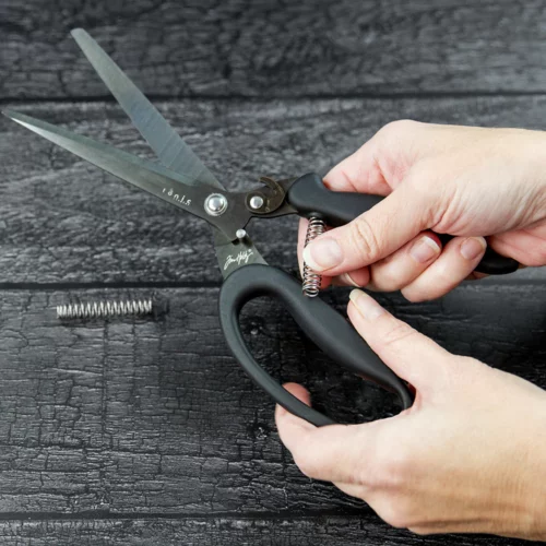 A persons hand can be seen coming out of the right hand side of the frame holding a pair of Tim Holtz Recoil Scissors. They are showing the spring mechanism of the snips. There is a faux wooden background.