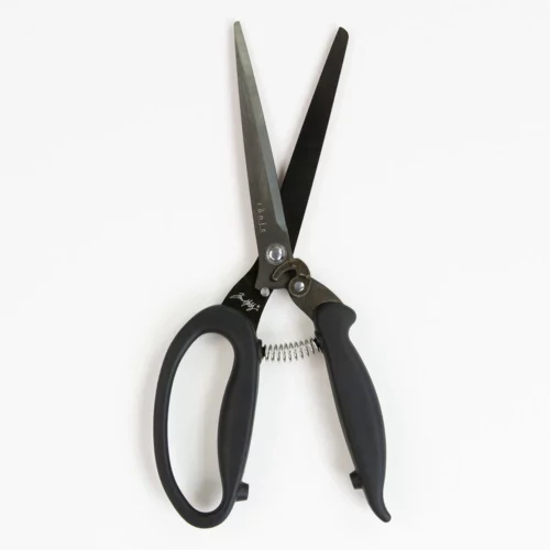 A single pair of Tim Holtz Recoil Scissors can be seen laying vertically in the center of the frame. The scissors have a black plastic handle and a metal blade with a spring between the handles. On a light grey background.