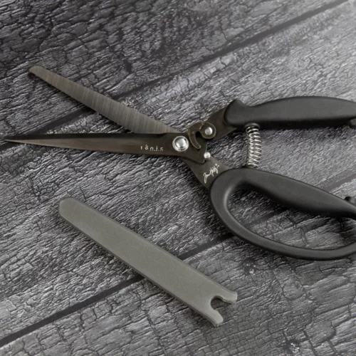 A pair of Tim Holtz Recoil Scissors can be seen laying on a faux wooden background. The snips have a black plastic handle and a metal blade with a spring between the handles. Laying on a faux wooden background.