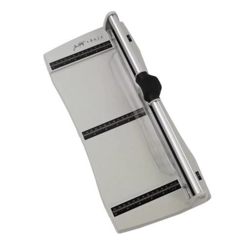 The Tim Holtz Rotary Media Trimmer has a light grey plastic base with grid marks debossed on the surface. It has a imperial and metric rulers with an extendable base and a geared rotary trimmer arm. The trimmer is rectangular in shape and is shown in the frame at a slight angle on a white background.