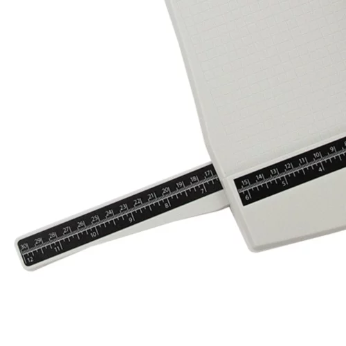 The Tim Holtz Rotary Media Trimmer has a light grey plastic base with grid marks debossed on the surface. It has a imperial and metric rulers with an extendable base and a geared rotary trimmer arm. In this frame, a close up of the corner of the trimmer is shown coming in from the top right hand side of the frame with one of the extendable rulers shown coming out of the trimmer. The image is on a white background.