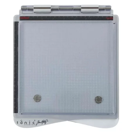 The Tim Holtz Stamping Platform is shown in the center of the frame. It is a birds eye view. The platform has a grey base and a clear perspex lid that is hinged at the top of the frame. On a white background.