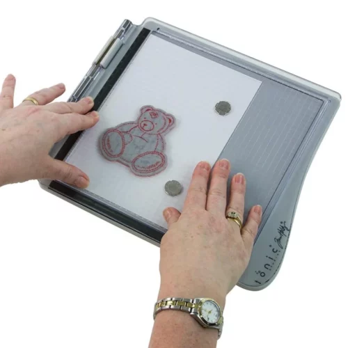 A persons hands are seen coming out of the bottom and left hand side of the frame, holding a Tim Holtz Stamping Platform. The platform is closed. The hands are pressing the clear perspex lid down with a stamp on the inside of the lid that is printing the stamp onto a piece of paper that is held in position using magnets on the bottom of the stamping platform. On a white background.