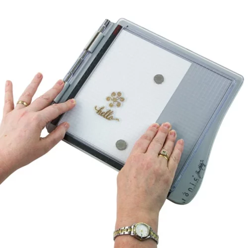 A persons hands are seen coming out of the bottom and left hand side of the frame, holding a Tim Holtz Stamping Platform. The platform is closed. The hands are pressing the clear perspex lid down with a stamp on the inside of the lid that is printing the stamp onto a piece of paper that is held in position using magnets on the bottom of the stamping platform. On a white background.