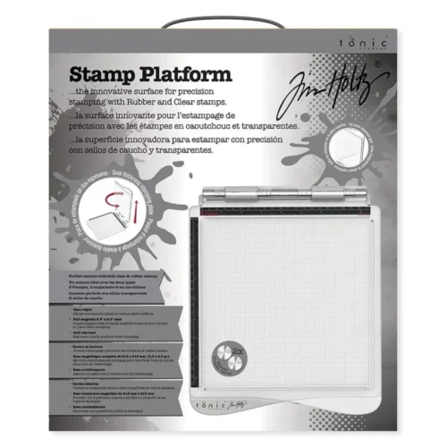 A box containing a Tim Holtz Stamping Platform is shown in the center of the frame. The box is square and has a picture of the stamping platform on the box with text describing the contents of the box. On a white background.