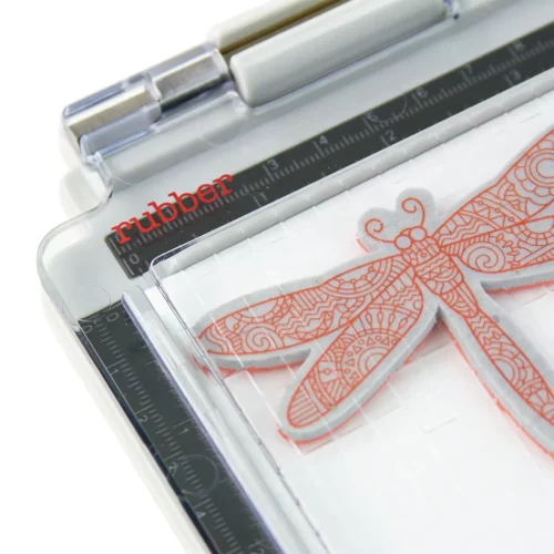 A close up of the a corner of the Tim Holtz Stamping Platform. The clear perspex lid is closed and there is a red word that indicates what stamp can be used on this side of the perspex lid. On a white background