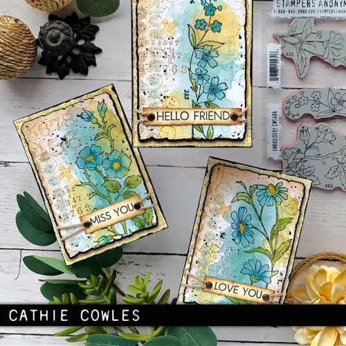 Three cards that were made using the Embroidery Tim Holtz Stamp Set are shown in the frame, with greenery scattered around them. The cards are stamped with flowers and are coloured in with blues and yellows and each card has a sentiment on it. The image is cut off by the frame.