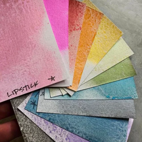 Some pieces of paper are fanned out in the frame. They have been sprayed with the different colours of Tim Holtz Distress Spritz. They are samples of the colours available in the range.