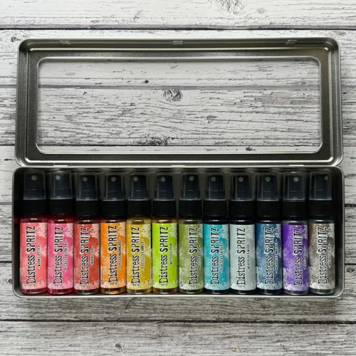 There is a storage metal tin shown in the center of the frame, with the lid open and you can see 12 Tim Holtz Distress Spritzes inside the tin. The bottles are a clear plastic with a black plastic spray top and a clear plastic lid. There is a coloured label around the body of each bottle that indicates the colour of the ink. The Distress Logo and product details are printed on the label. The tin is laying on a faux wooden background.