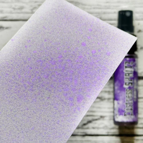 A sample of the Wilted Violet Tim Holtz Distress Spritz is shown on a piece of cardstock that is seen coming in from the left hand side of the frame. The Distress Spritz bottle is laying in the background on a faux wooden background and the bottle is blurred so that the focus is on the paper and the sample of the contents of the bottle.