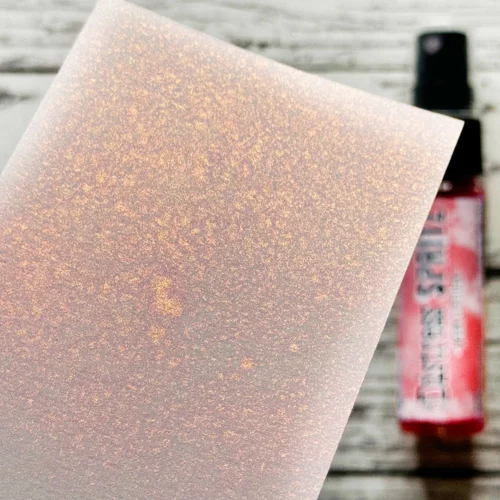 A sample of the Worn Lipstick Tim Holtz Distress Spritz is shown on a piece of cardstock that is seen coming in from the left hand side of the frame. The Distress Spritz bottle is laying in the background on a faux wooden background and the bottle is blurred so that the focus is on the paper and the sample of the contents of the bottle.