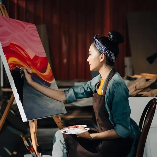 a woman is sitting in a chair in a room with wood panel walls. she is wearing a green jersey and a brown apron with a blue flowered headband. she is holding a palette that has paint on it in her left hand and a paintbrush in her right hand. she is leaning forward and painting on a large canvas that is standing on an easel. the canvas has a picture of a sunset on it. the colours are pinks, pranges and reds. it is in the centre of the image.