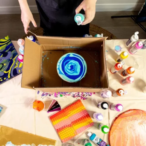 there is a cardboard box in the image and it has a round canvas inside of it that is being poured on with the acrylic paint. in shades of blue and white. there is a person pouring paint onto it wearing a black apron. across the table there are various bottles of different colours all visable from a birds eye view. infront of the person you can see the other canvases they were working on in various colours. in the centre of the image.