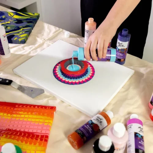 at the centre of the image the mont marte acrylic pouring paint is being used to pour a light blue colour onto the centre of a canvas on a table. these are other colours in rings that have been poured before. the other bottles of the mont marte acrylic pouring paint are laid out on the table with their labels facing upwards. on the left hand side of the canvas is a premade canvas with orange and yellow and red colours on it. they are resting on a light tablecloth. a persons hand can be seen pouring the paint and they are dressed in black