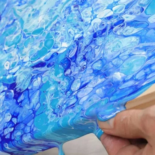 Mont Marte Premium Silicone Oil is shown being used on a canvas. the canvas is slighlty tilted towards the bottom of the screen. the colours on the canvas are shades of light blue and white with a touch of dark blue. there are cells in the paint of varoius sizes and the paint is running off of the canvas. the bottom right corner is a persons hands holding and tilting the canvas. in the center of the image