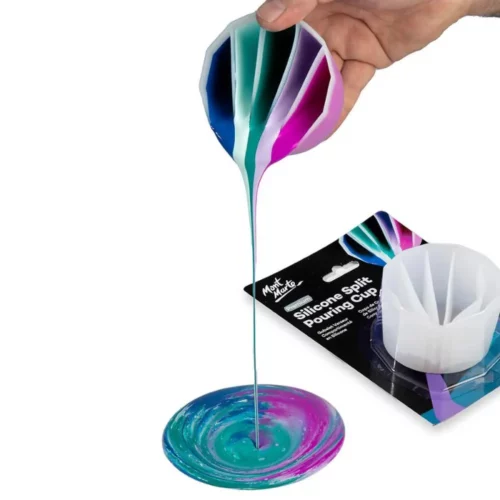 Mont Marte Silicone Split Pouring Cup is shown in use at the top of the image. there are shades of pink, blue and black inside each section of the cup and it is being tilted so that the paint is pouring out of it onto a circle patch on the table where the colours are creating patterns as they fall into eachother. behind this is a picture od the cup in its original packaging. in the centre of the image on a white background.