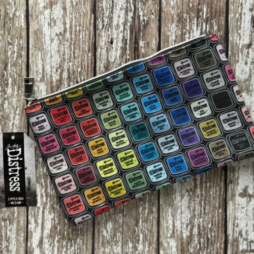 A Tim Holtz Distress Zipper Bag Medium is shown diagonally in the center of the frame. The bag is made of a printed fabric, adorned with colorful artwork from the complete Distress colour palette. The bag has a Distress tag attached to it and it has a metal zipper. The bag is laying on a faux wooden background.