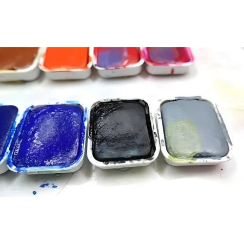 in the front there are three gouache colours lined up from left to right, blue black and grey. behind then you can see the tops of the other colors of pinks and orange and reds. they are all on a white table surface