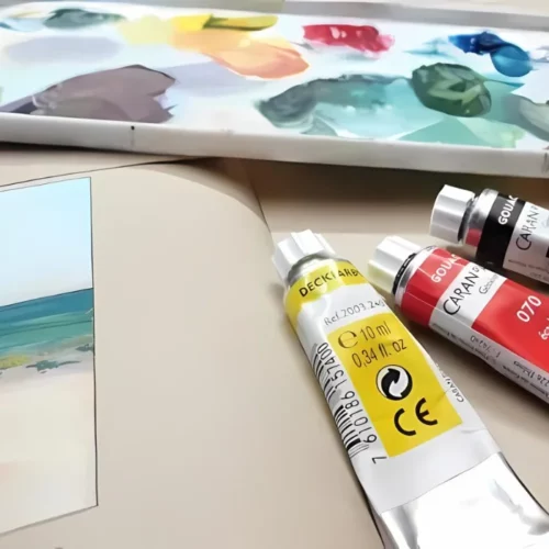 Three tubes of Caran D'Ache Studio Gouache Paint are seen laying on a pad with a plastic pallete covered in ink, in the background, which is slightly blurred.