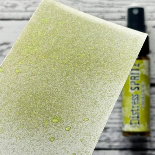 A sample of the Crushed Olive Tim Holtz Distress Spritz is shown on a piece of cardstock that is seen coming in from the left hand side of the frame. The Distress Spritz bottle is laying in the background on a faux wooden background and the bottle is blurred so that the focus is on the paper and the sample of the contents of the bottle.