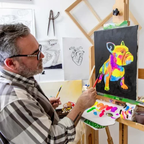 A man is seen painting an image of a dog using the Fluoro Mont Marte Premium Acrylic Paint Set. The dog is fluorescent and multi coloured.